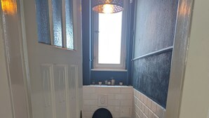 A narrow bathroom with a toilet, a window with a decorative lamp, and a patterned floor. - Cozy old building with sauna and view (Wuppertal)