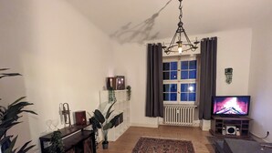 A living room with a wooden floor, a large rug, a TV, a radiator, and a window with curtains. - Cozy old building with sauna and view (Wuppertal)