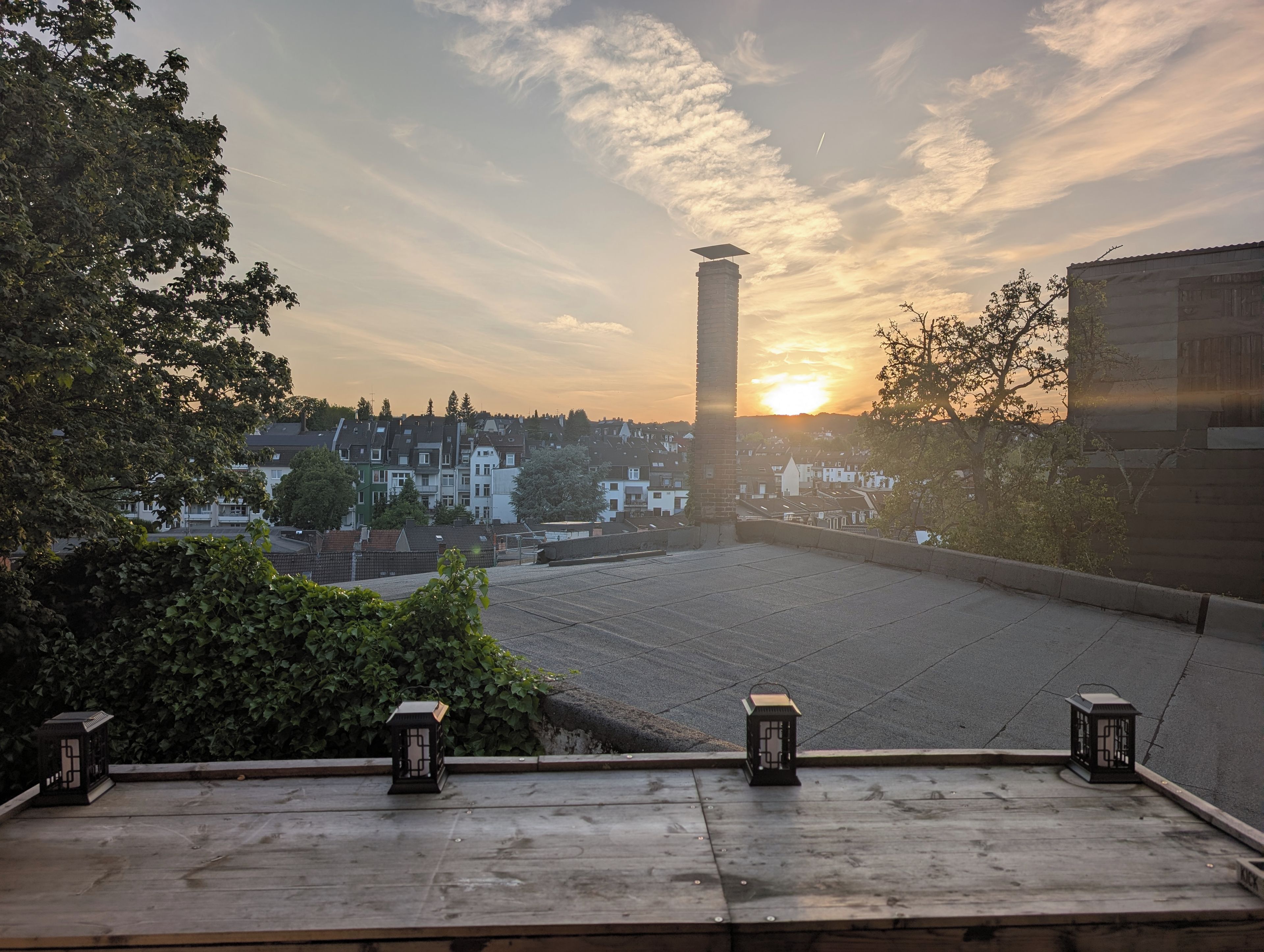 A rooftop terrace with a view of a sunset over a residential area, including a tall chimney and a row of houses.