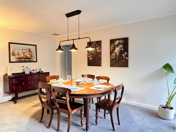 Un comedor con una mesa redonda de madera, seis sillas, dos lámparas colgantes, un mueble de madera oscura y dos cuadros en la pared.