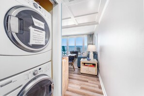 A laundry area with stacked washers and dryers, a view of the balcony, and a seating area with a lamp and books. - 5th Floor Oceanfront Condo w/ Outdoor Pool by Seaside Vacations & Sales (Ocean City)