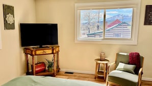 Een kamer met een bed, een televisie op een houten standaard, een stoel, een tafeltje en een raam met uitzicht op een huis en bergen.