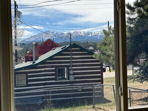 Une cabane en rondins avec une clôture en métal à l’avant, un bâtiment à toit rouge en arrière-plan et des montagnes enneigées.