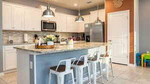 Uma cozinha moderna com armários brancos, uma ilha central com banquetas, eletrodomésticos de aço inoxidável e iluminação pendente.