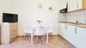 Ett kompakt kök med vita skåp, ett matbord med stolar, en tv och en liten krukväxt.