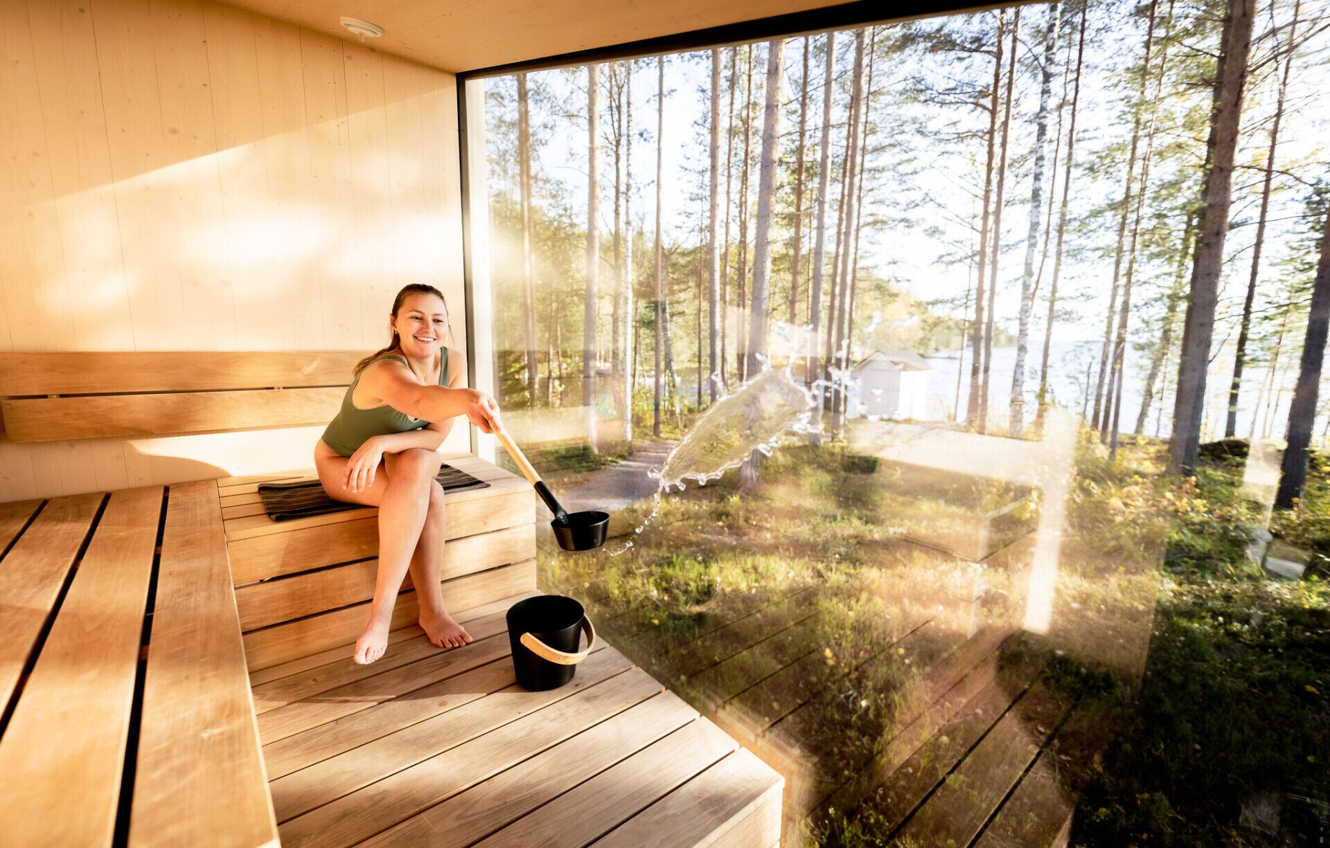 A person sitting on a wooden bench in a sauna area with a view of a forest and a body of water.
