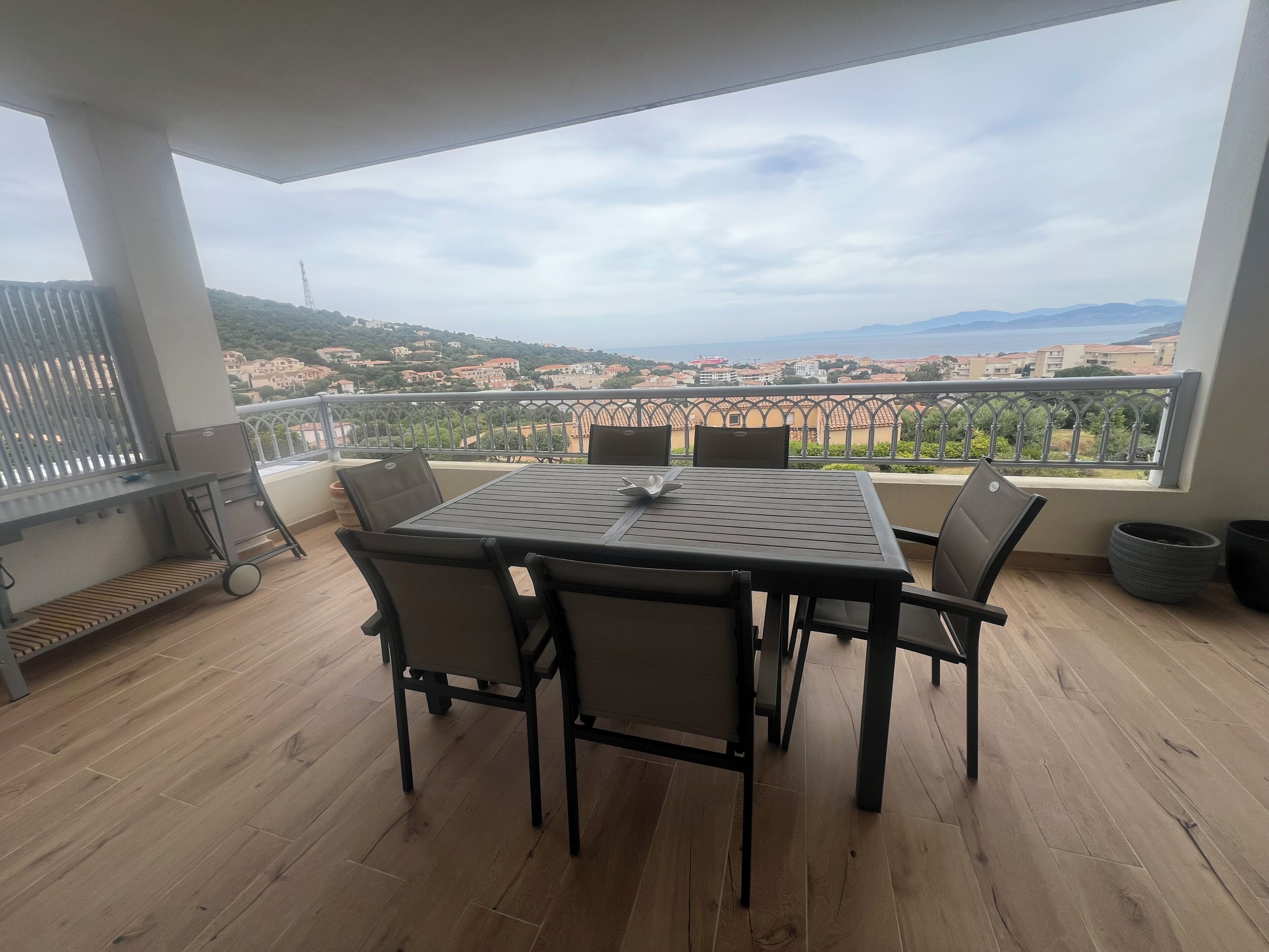 Een balkon met een tafel, stoelen, een barbecue en uitzicht op een kustplaatsje.