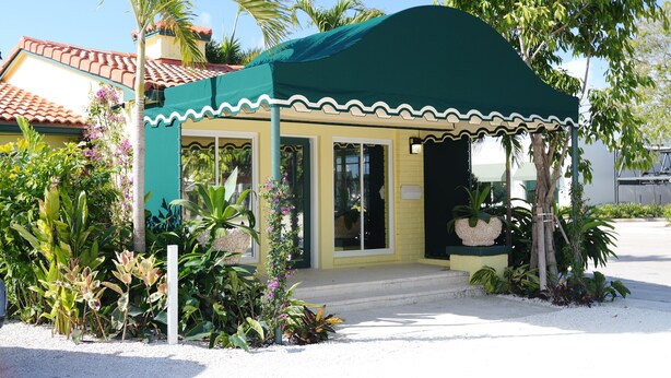 A small, single-story building with a green awning, a tiled roof, and a garden with various plants and flowers.