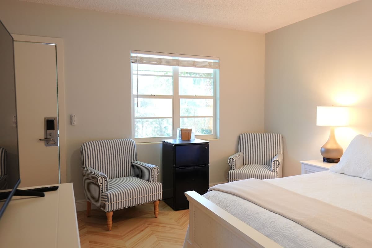 Ein Hotelzimmer mit einem Bett, zwei gestreiften Sesseln, einem kleinen Tisch mit einem Laptop, einem Fenster mit Jalousien und einer Nachttischlampe.