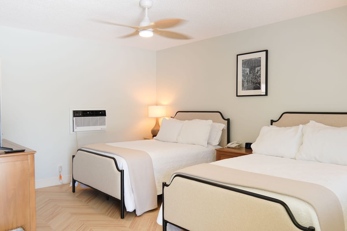 Ein Hotelzimmer mit zwei Betten, einem Schreibtisch mit Computer, einem Deckenventilator, einer Wandheizung, einer Nachttischlampe und einem gerahmten Bild an der Wand.