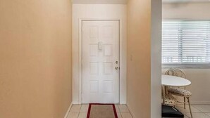 Un pasillo con una puerta blanca, un pequeño comedor con una mesa y sillas, y una ventana con persianas.