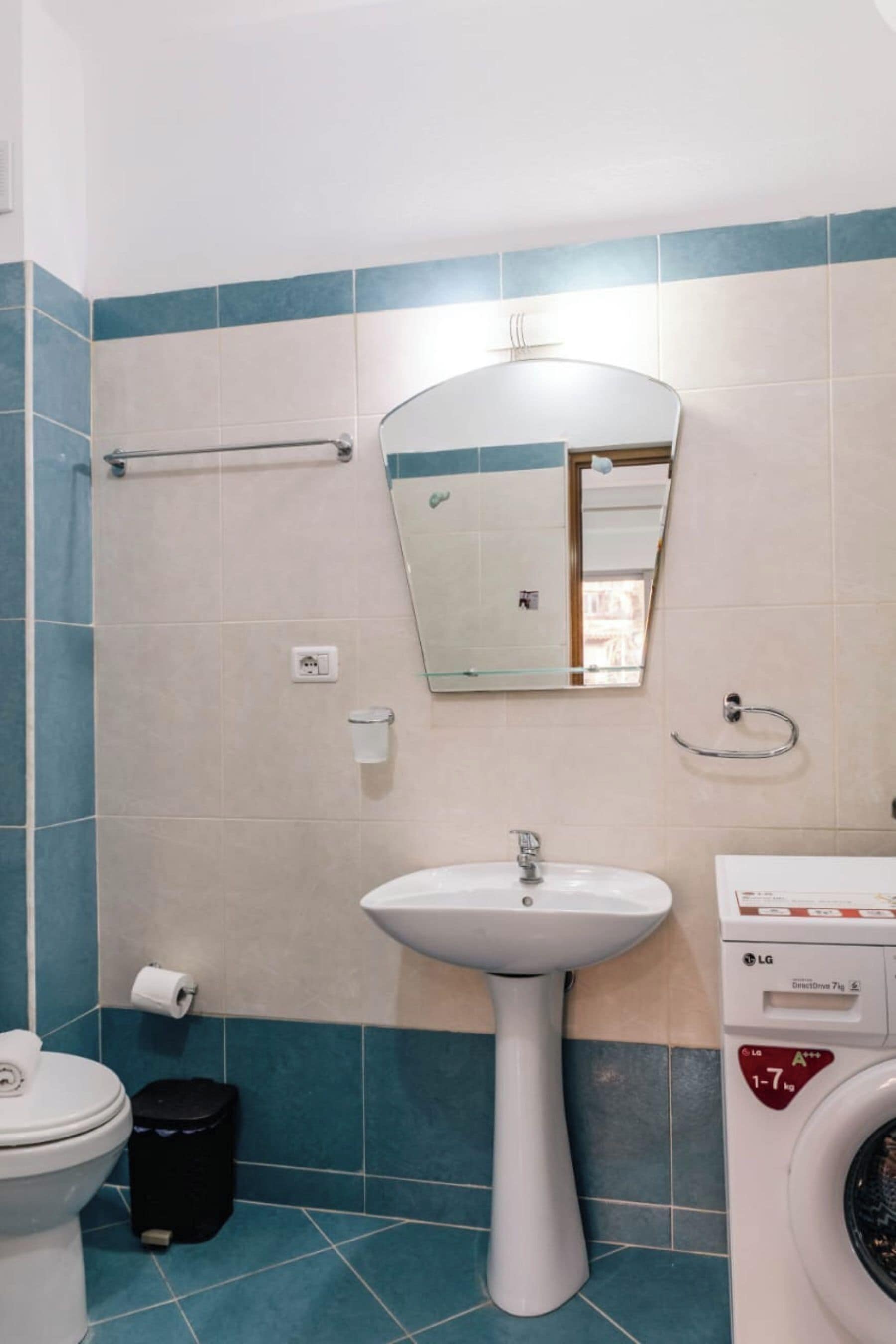 Een badkamer met een wastafel op voetstuk, een spiegel, een handdoekhouder, een toilet, een prullenbak en een wasmachine.