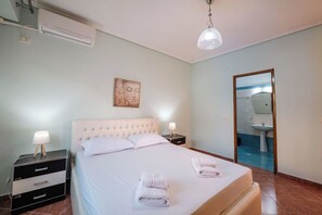 Un dormitorio con cama, mesitas de noche, aire acondicionado de pared, lámpara de techo y baño visible a través de una puerta abierta.