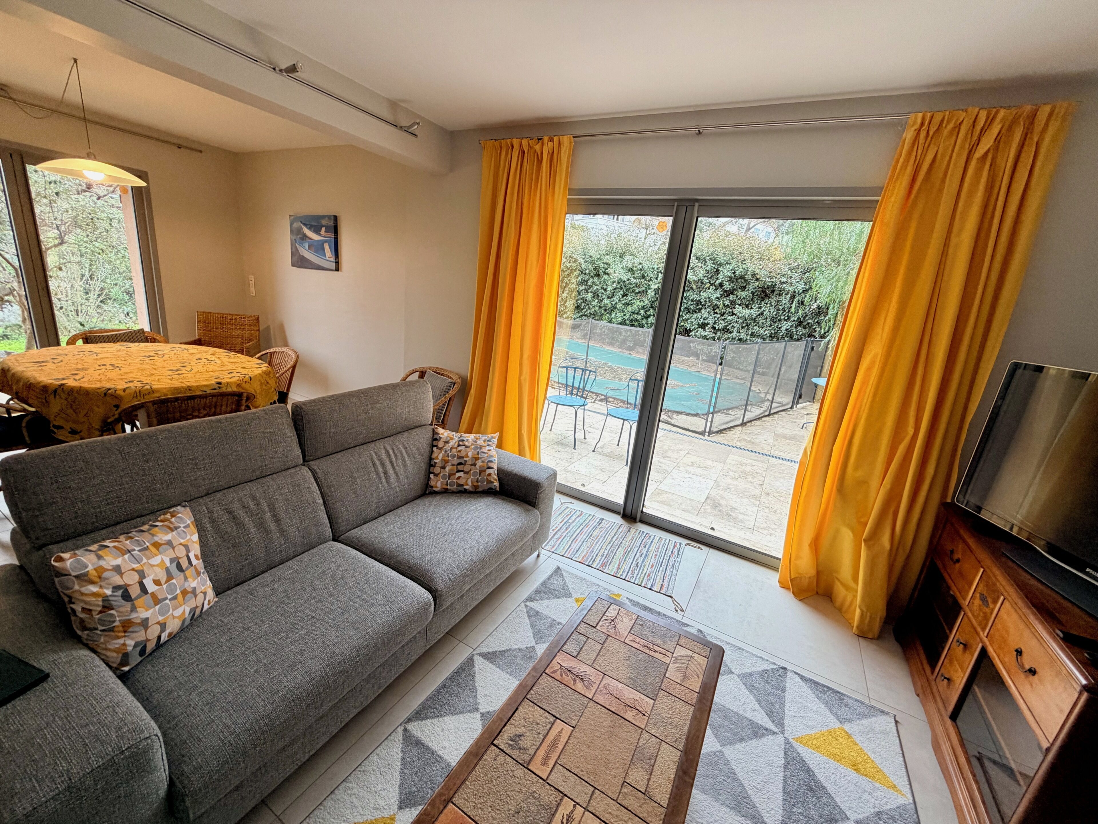 Sala de estar com sofá, televisão, mesa de jantar e cadeiras, e vista para área externa através de uma porta de vidro.