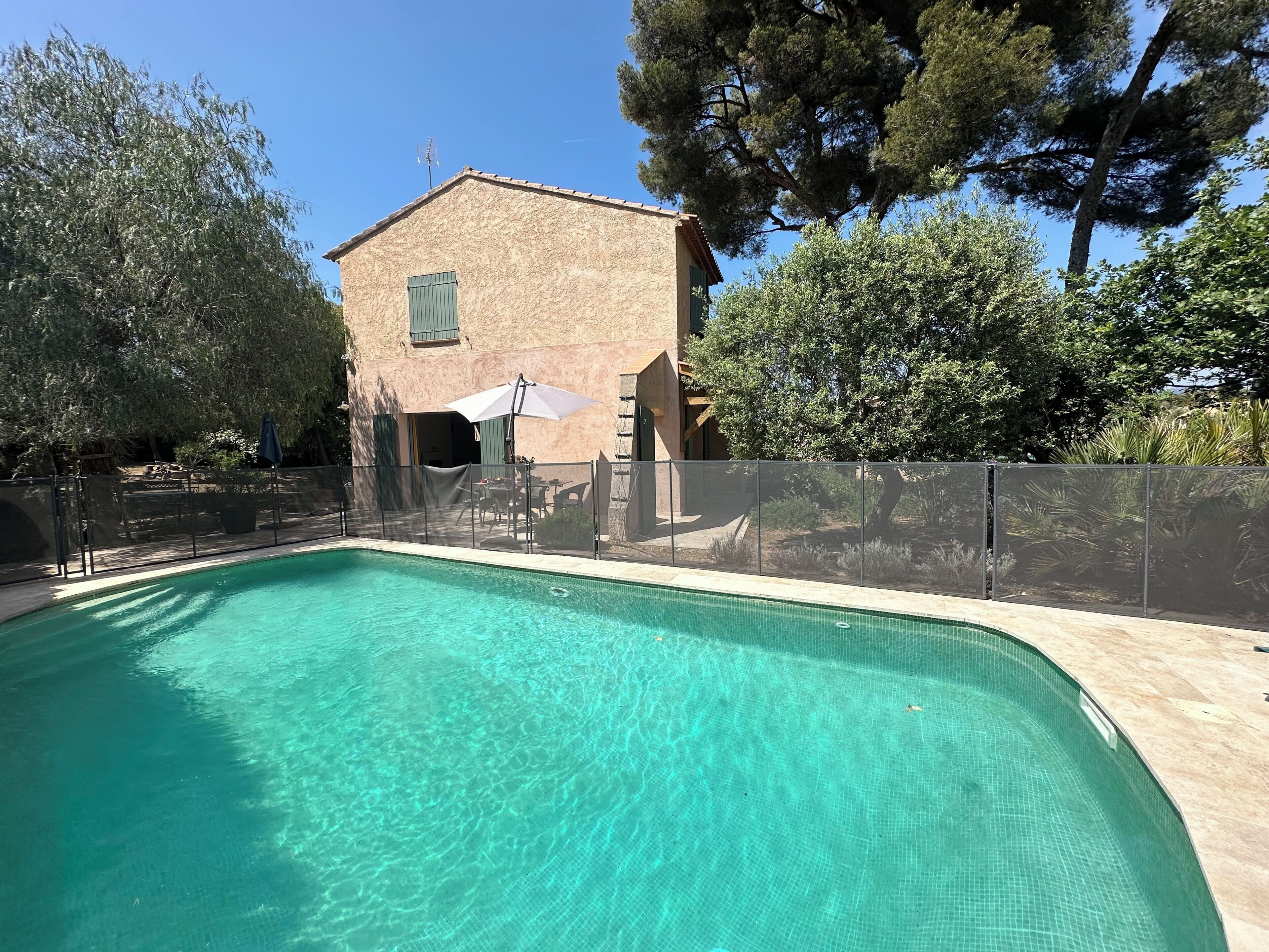 Una piscina con agua cristalina azul, rodeada por una cerca, árboles y un edificio con tejado de baldosas.