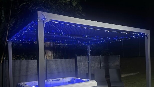 A hot tub under a covered patio with string lights, surrounded by a fence and outdoor seating. - TROPICAL BUNGALOW (Gros-Morne)