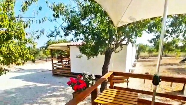 A wooden deck with a bench, a small table, and a white building in the background. - Feudo Mallav b&b Noto (Avola)