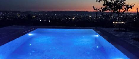 A rectangular pool with blue lighting, surrounded by a cityscape at dusk.
