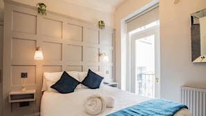 A hotel room with a bed, bedside tables, a nightstand, a blue throw blanket, a white headboard, wall-mounted lights, and a window with a view. - Second Floor Suite | Rathmines | WiFi & Comfort (Dublin)