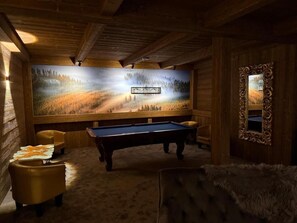 Una mesa de billar en una habitación con paredes de madera y un cuadro grande de un paisaje forestal.