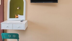Una habitación con una televisión montada en la pared, un pequeño mueble blanco con cajones, una silla verde y un espejo con luz integrado en la pared.