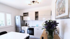 Een moderne keuken met witte kastjes, een roestvrijstalen koelkast, een vaatwasser en een koffiezetapparaat. Er staat een eettafel met stoelen en op de zijkant een vaas met bloemen.