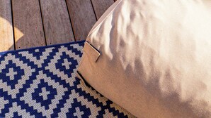 Une terrasse en bois avec une piscine, un coussin beige et un tapis à motifs.