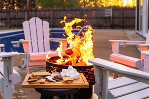 A fire pit with flames, surrounded by Adirondack chairs, a pool, and a table with snacks.