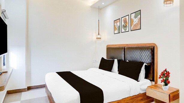 A modern hotel room with a large bed, a wooden headboard, a bedside table with a vase of flowers, and a wall-mounted light fixture. - Deluxehotel Surabhi Elite (Hyderabad)
