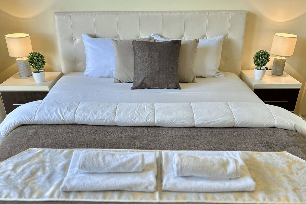Una cama bien hecha con edredón gris y blanco, dos lámparas de noche y una pequeña planta en maceta sobre cada mesita.