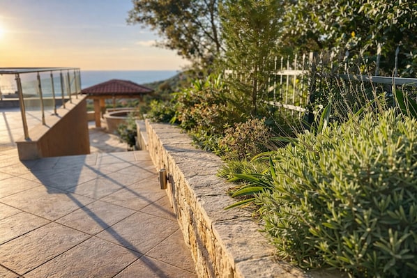 Un chemin de pierre mène à une balustrade en verre qui surplombe une étendue d’eau, avec une petite construction et une végétation luxuriante.