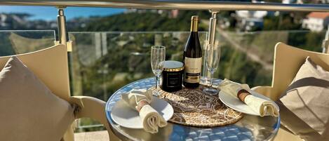 A table with a bottle of wine, two glasses, and a small container on a balcony overlooking a scenic view.