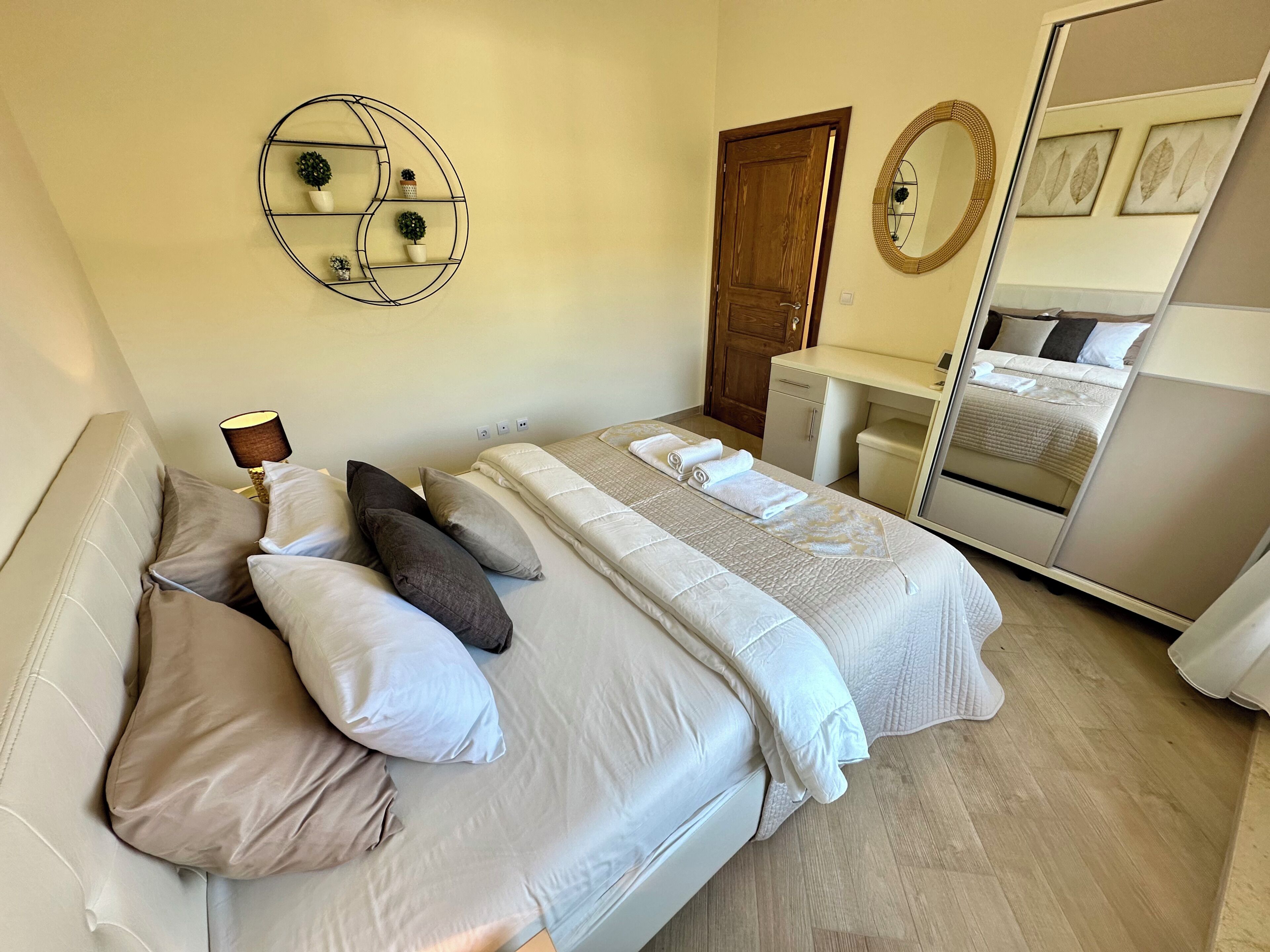 A bedroom with a bed, pillows, a bedside table, a mirror, and a wall-mounted shelf with decorative items.