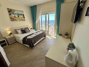 Ein Hotelzimmer mit Bett, Nachttisch, Lampe und Blick auf das Meer durch ein großes Fenster.