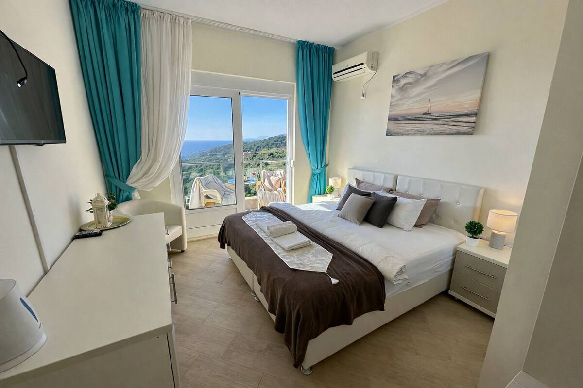 Una habitación de hotel con cama, televisor, ventana con cortinas, un cuadro y un equipo de aire acondicionado.