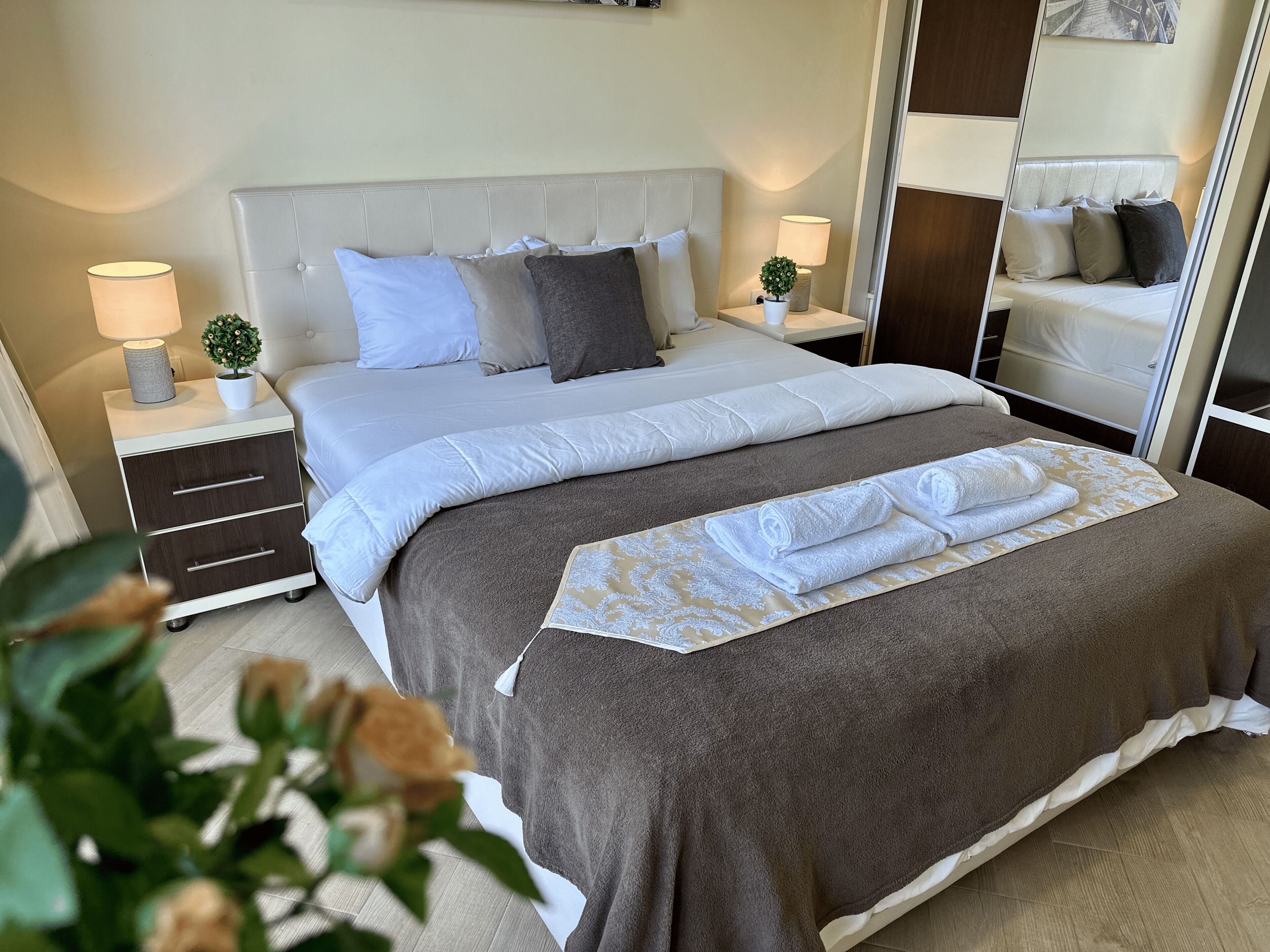 A neatly made bed with a brown blanket and white linens, two bedside tables with lamps and small potted plants, and a large mirror on the wall.