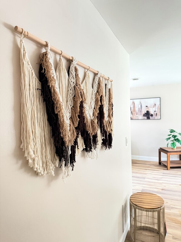 Une décoration murale en macramé aux couleurs variées, un tableau encadré accroché au mur et une petite table ronde sur laquelle est posée une plante.