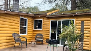 A log cabin with a deck, wicker chairs, a grill, and a potted plant.