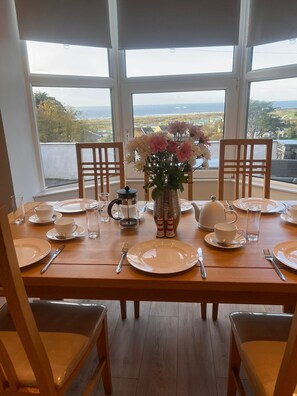 A dining table set for four with chairs, a vase of flowers, and a view of the sea through large windows.
