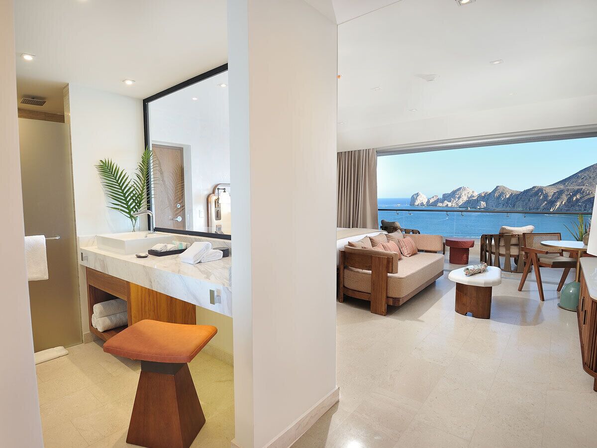 Habitación de hotel moderna con baño que incluye un espejo grande, un lavamanos y tocador. La zona de estar cuenta con un sofá, una mesa de centro y vistas al mar.