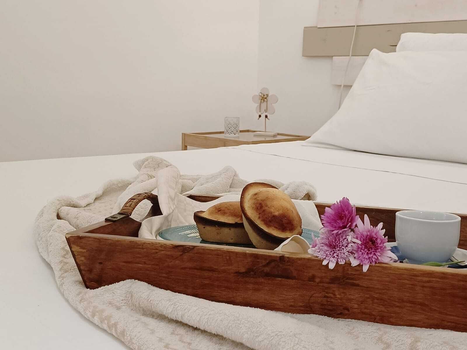 A neatly made bed with white linens, a wooden tray with bread and flowers, and a bedside table with a lamp and a small decorative item.