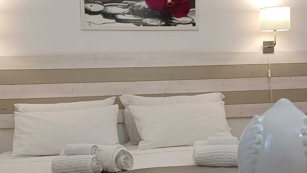 Habitación de hotel con cama, almohadas, una toalla enrollada, una lámpara de pared y un cuadro enmarcado.