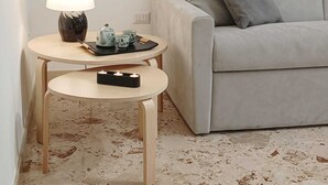 Un salón con un sofá gris, una mesita redonda con una lámpara, un hervidor y tazas, además de un pequeño taburete.