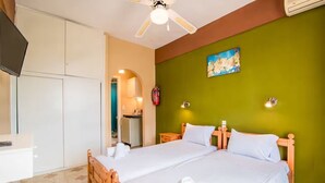 Quarto de hotel com cama, escrivaninha, televisão, ventoinha no teto, área de cozinha e extintor de incêndio.