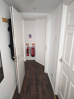 A hallway with white walls, a wooden floor, and two fire extinguishers mounted on the wall. - Duplex Apartment in the Heart of Glenarm Village (Ballymena)