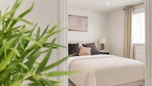 Quarto com cama, mesa de cabeceira, luminária, janela com cortinas e uma planta.