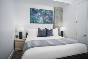 Una cama bien hecha con una colcha gris, dos lámparas de noche, una planta pequeña y un cuadro en la pared.