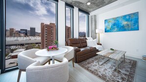 Een moderne woonkamer met uitzicht op de stad, een bruine leren bank, een ronde witte tafel en een blauw abstract schilderij.