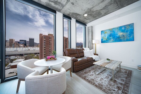 Una sala de estar moderna con vista a la ciudad, un sofá de cuero marrón, una mesa redonda blanca y un cuadro abstracto azul.