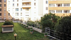 Un edifici residencial de diversos pisos amb balcons, una zona verda amb plantes en testos i una escala metàl·lica.